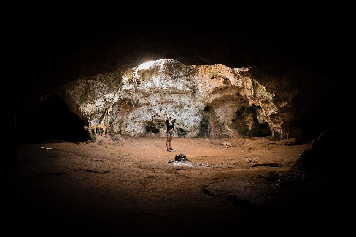 A visitor standing inside a large sunlit cave chamber lit by a natural ceiling opening, like Aruba's Quadirikiri Cave
